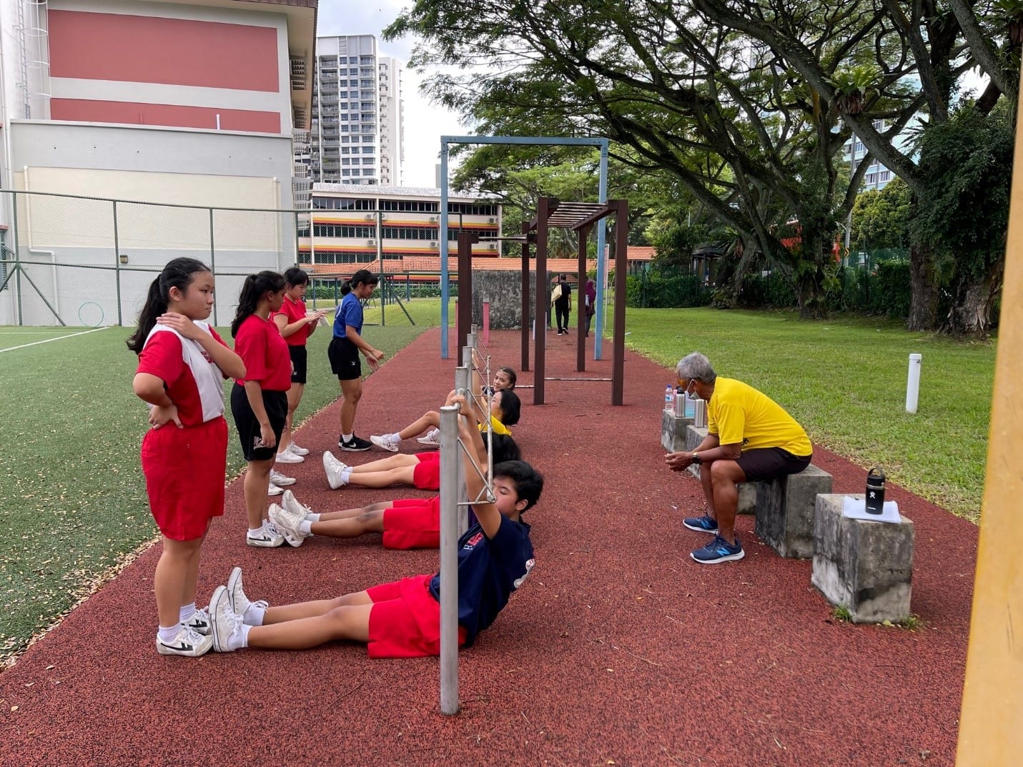 Students in red uniforms exercise on a red track as an older man in yellow watches, with trees and buildings behind.